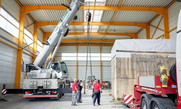A large wooden crate is lifted off a trailer by crane in a hall.