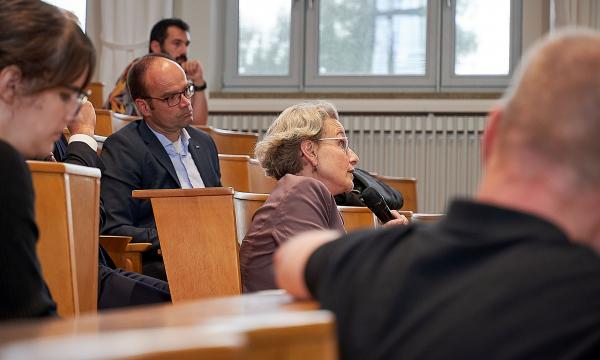A lecture hall, a woman with a microphone asks a question. A man with glasses sits behind her and listens.
