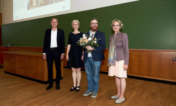 2 women and 2 men stand next to each other in front of the blackboard in a lecture hall. One man holds a bouquet of flowers.