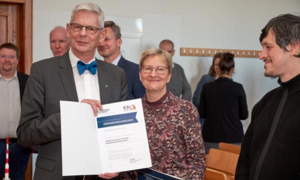 älterer Mann und Frau mit Brille lächeln in die Kamera, er hält eine Urkunde
