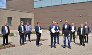 10 men in suits stand in front of a building. Two are holding a document in their hands.