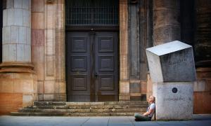 Ein Mann sitz am Boden, angelehnt an große Granitsteine vor einer Holztür.