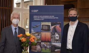 Farewell to Prof. Günter Löffler (l.) by the Dean of the "Friedrich List" Faculty of Transport and Traffic Sciences, Prof. Günther Prokop Two men with face masks next to each other, one holding flowers, a rollup in the middle.