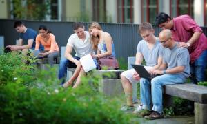 Das Foto zeigt sieben Studierende, die in kleinen Grüppchen auf Bänken im Freien sitzen und lernen.