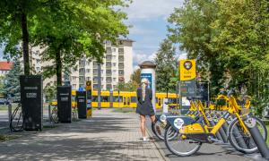 A tram in the background, e-bikes parked on the right, charging stations on the left, a woman