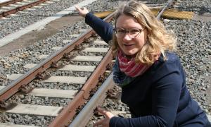 A woman with blond hair and glasses squats by a railway track and points at something.