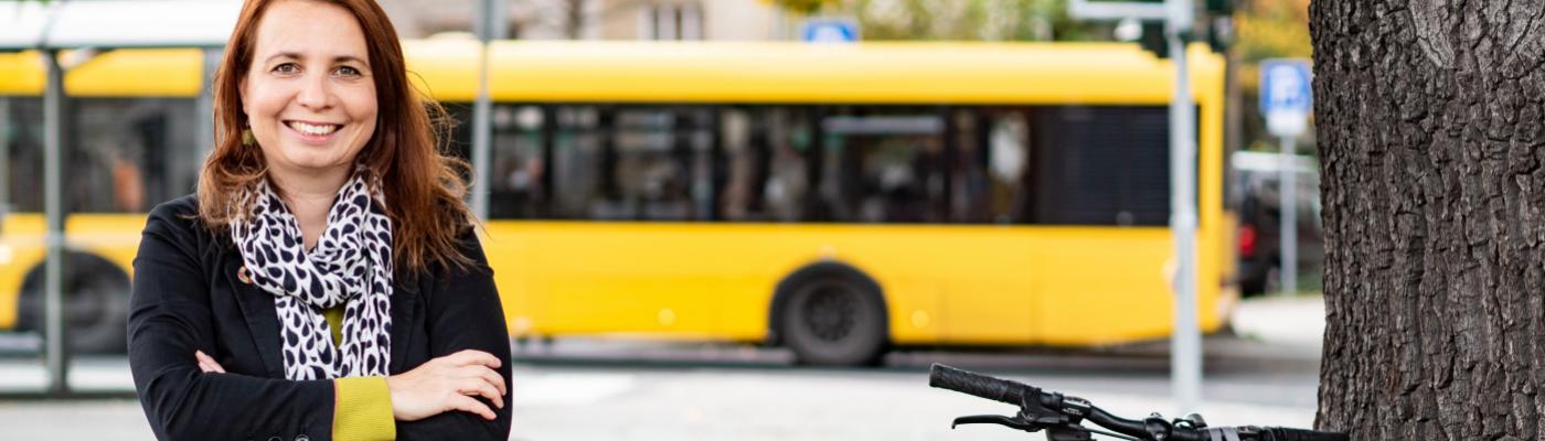 Eine Frau steht mit verschränkten Armen am Straßenrand, hinter ihr fährt ein Bus.