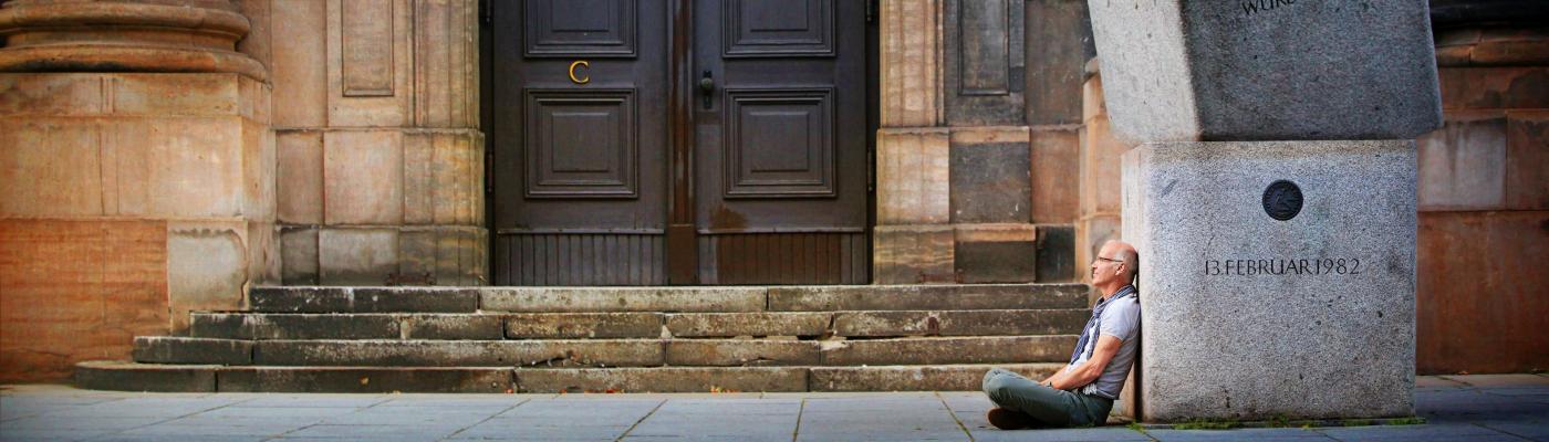 A man sits on the floor, leaning against large granite stones in front of a wooden door.