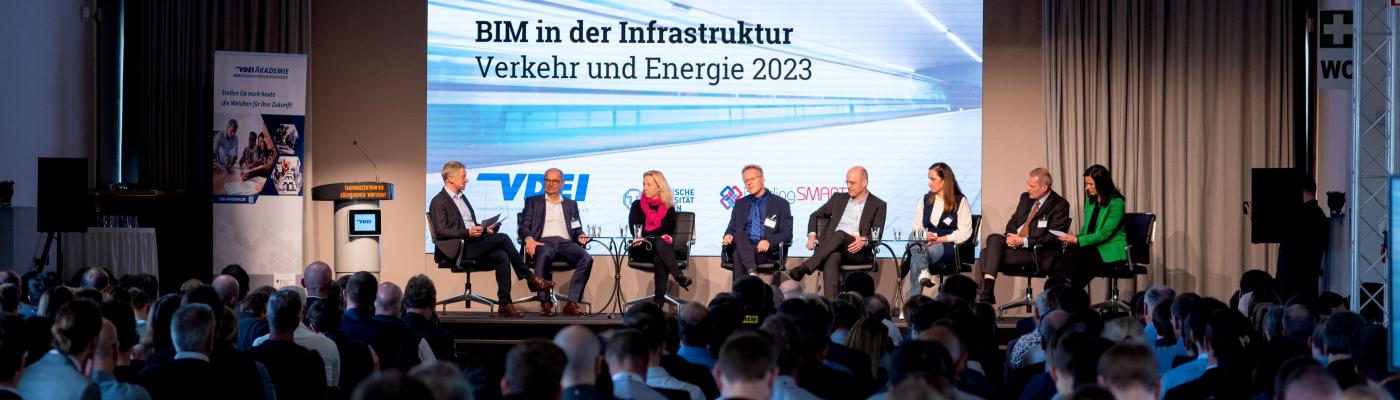 A panel discussion on a stage with 8 participants in front of many spectators