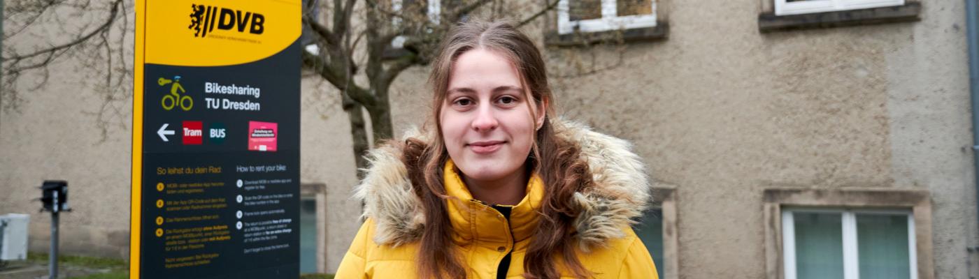 Eine junge Frau in einer gelben Jacke steht vor einem Schild und einem Gebäude