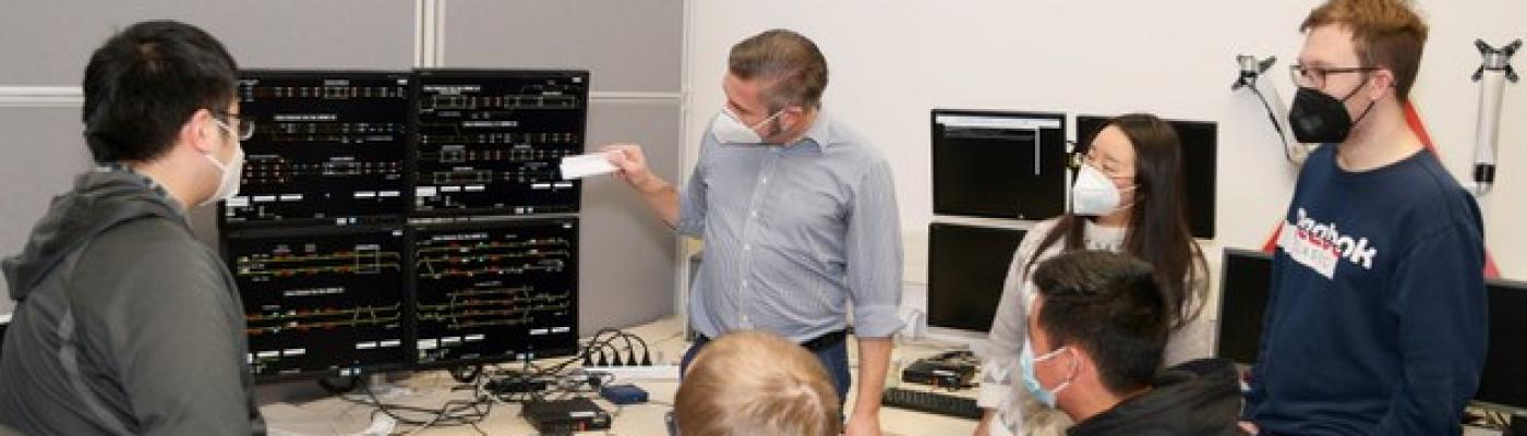 Seminar im Sicherungstechnischen Labor der TU Dresden. 6 Menschen stehen und sitzen vor einem technischen Gerät.