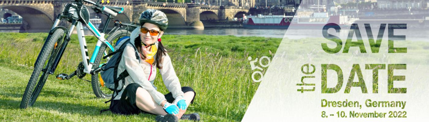 A cyclist sits on a meadow, her bike next to her, the old town of Dresden in the background.