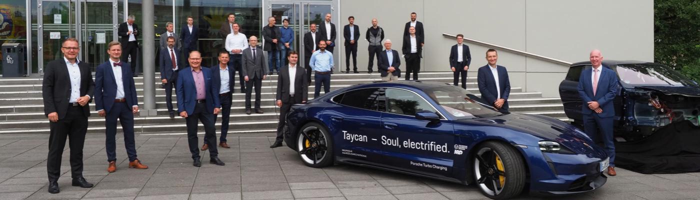 Ein Gruppe von Menschen steht vor einem Gebäude, dazu ein Porsche und ein Auto-Cockpit.