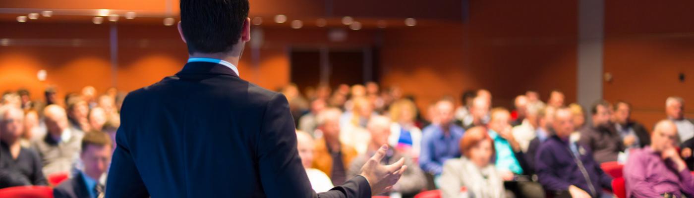 The photo shows a speaker giving a presentation to a large group.