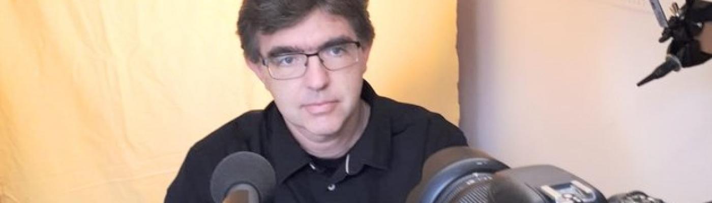 Ein Mann sitzt hinter einem Mikrofon und Laptop. Er trägt eine Brille und ein dunkles Hemd.