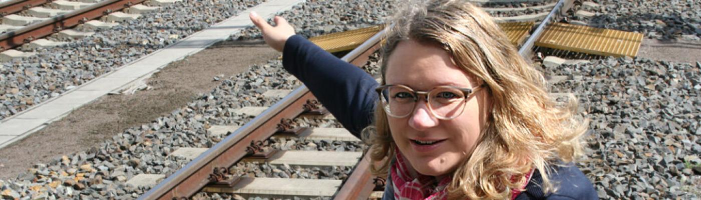 A woman with blond hair and glasses squats by a railway track and points at something.